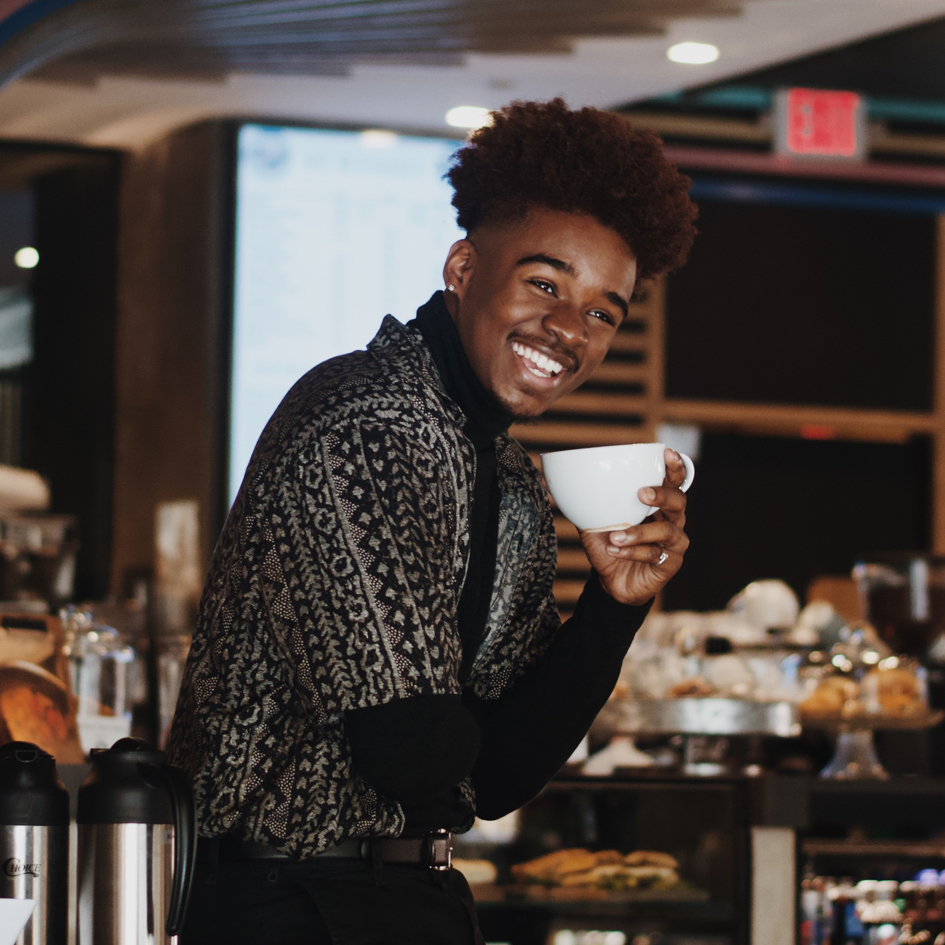 Smiling person with a patterned shirt drinking coffee infront of a baked goods display