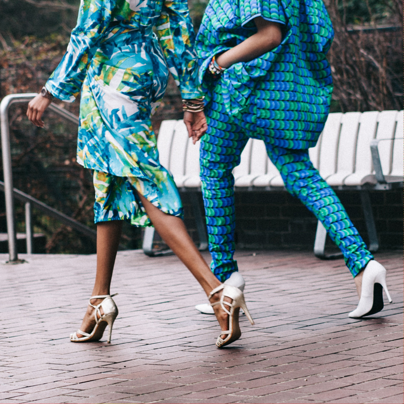 Two models in white heels walking outside by a park bench in turqouise outfits